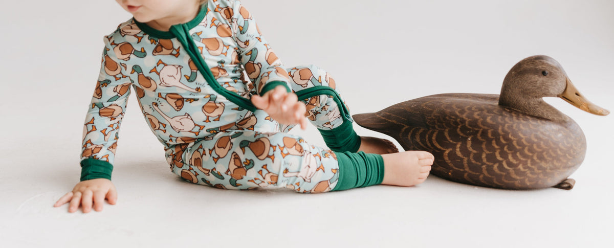 Child wearing a pair of zip-up pajamas with a pattern containing ducks and geese, sitting next to a wooden duck prop.