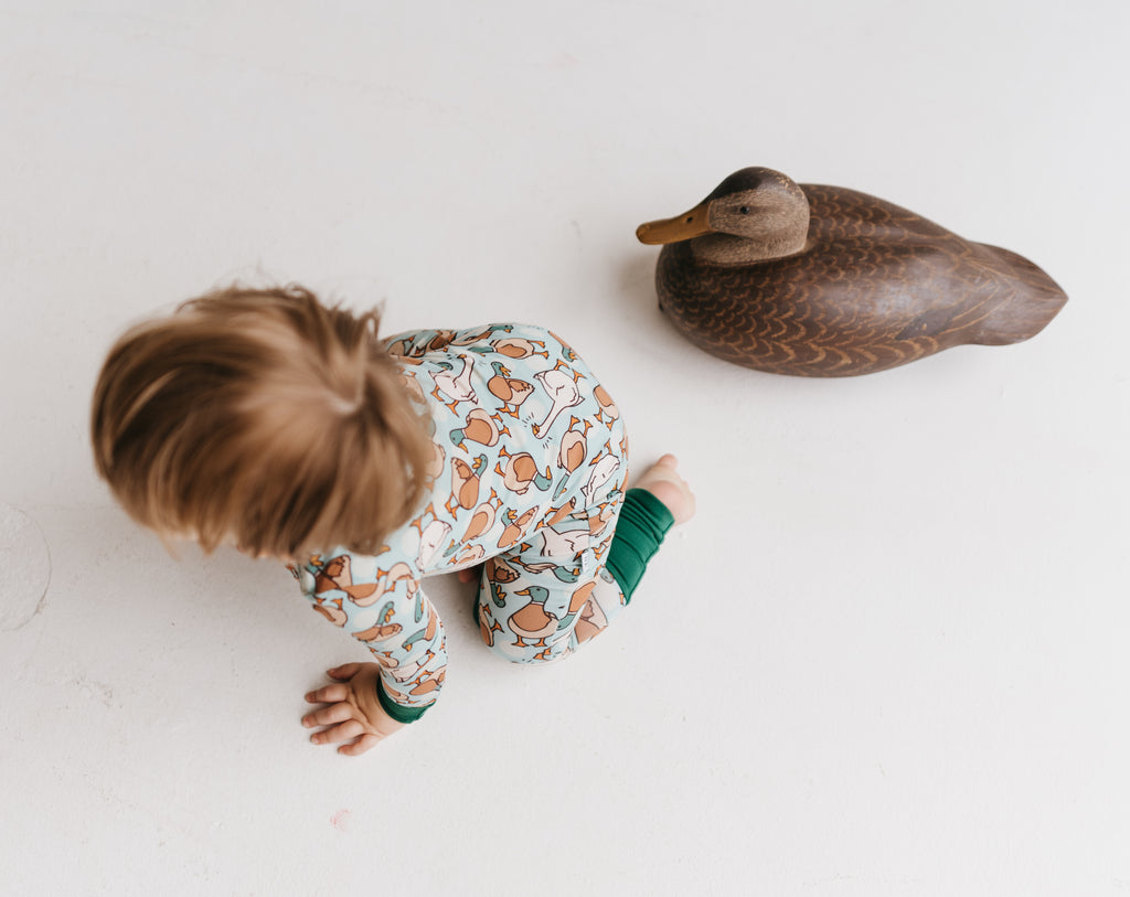 Child crawling on the floor next to a wooden duck toy in a pair of zip-up pajamas with a pattern containing ducks and geese.