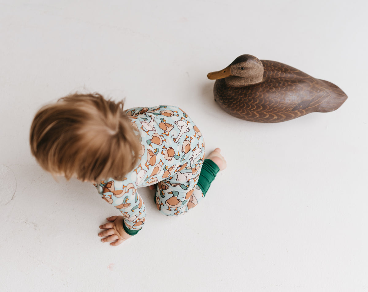 Child crawling on the floor next to a wooden duck toy in a pair of zip-up pajamas with a pattern containing ducks and geese.