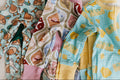 Three children's long-sleeve pajamas are laid flat on a light fabric surface. The left pajama has a duck print on a pale blue background, the middle has a patchwork pattern with pink, beige, and brown sections, and the right pajama is light blue with butter sticks and yellow hearts.