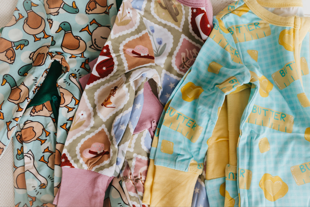 Three children's long-sleeve pajamas are laid flat on a light fabric surface. The left pajama has a duck print on a pale blue background, the middle has a patchwork pattern with pink, beige, and brown sections, and the right pajama is light blue with butter sticks and yellow hearts.