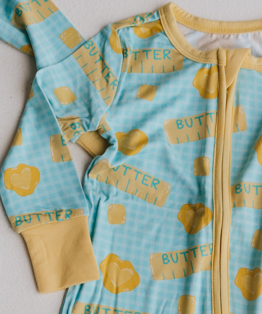 A baby long-sleeve zip-up sleeper in light blue with a gingham pattern and yellow butter-themed print, featuring yellow cuffs and a yellow zipper.