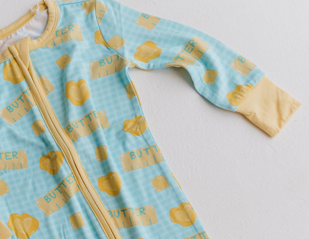 A baby long-sleeve zip-up sleeper in light blue with a gingham pattern and yellow butter-themed print, featuring yellow cuffs and a yellow zipper.