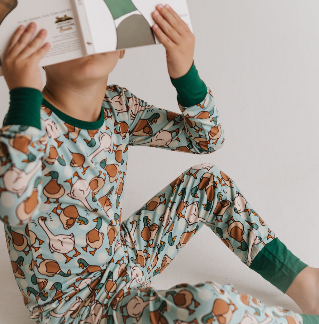 A child wearing a light blue long-sleeve pajama set with a brown and beige duck print and dark green cuffs, sitting and holding an open book over their face against a plain light background.