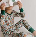 A child wearing a light blue long-sleeve pajama set with a brown and beige duck print and dark green cuffs, sitting and holding an open book over their face against a plain light background.