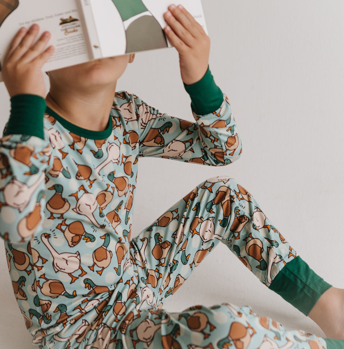A child wearing a light blue long-sleeve pajama set with a brown and beige duck print and dark green cuffs, sitting and holding an open book over their face against a plain light background.