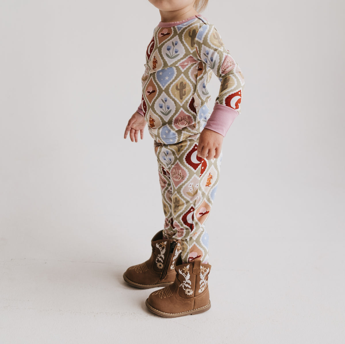 Toddler standing in profile wearing a long-sleeve matching pajama set with a western-themed argyle pattern in beige, brown, red, blue, and white, paired with brown western boots, against a plain white studio background.
