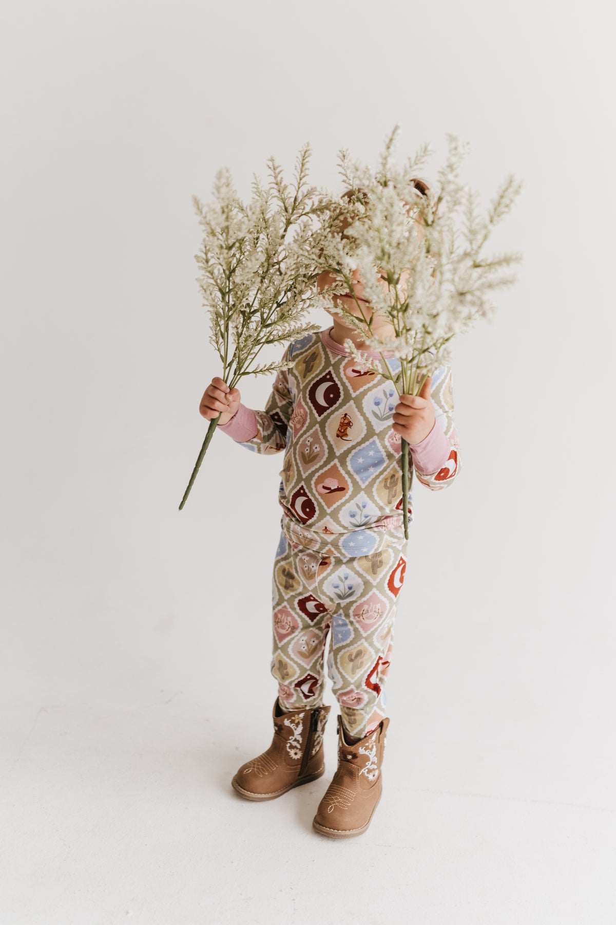 A toddler stands against a plain light background holding a large bouquet of white dried flowers. The child is wearing a one-piece jumpsuit with a multicolor retro geometric pattern in pink, blue, red, and beige, paired with brown cowgirl boots.