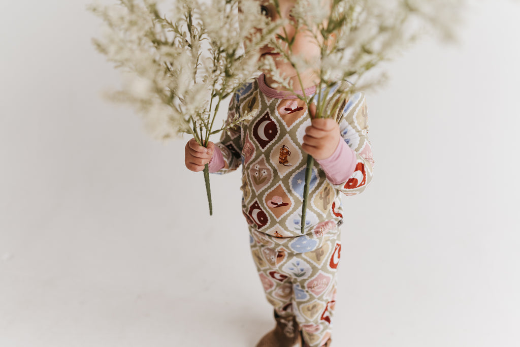 Toddler wearing a long-sleeve matching pajama set with a western-themed argyle pattern in beige, brown, red, blue, and white, paired with brown western boots holding white dried flowers in front of their face against a plain white background.