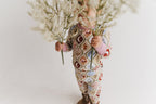 Toddler wearing a long-sleeve matching pajama set with a western-themed argyle pattern in beige, brown, red, blue, and white, paired with brown western boots holding white dried flowers in front of their face against a plain white background.