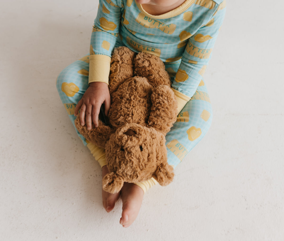 Child sitting down, holding a bear, wearing two piece pajamas with a yellow and blue butter pattern against a plain white backgroun.