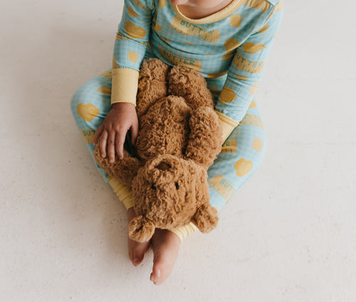 Child sitting down, holding a bear, wearing two piece pajamas with a yellow and blue butter pattern against a plain white backgroun.