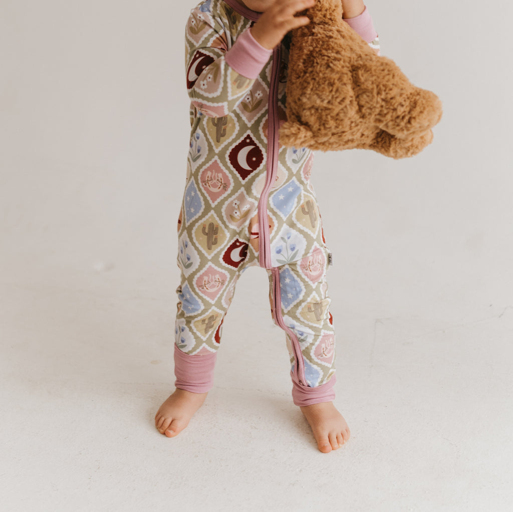 A baby wearing a pink country-themed patterned zip-up sleeper, holding a brown teddy bear against a plain off-white background.