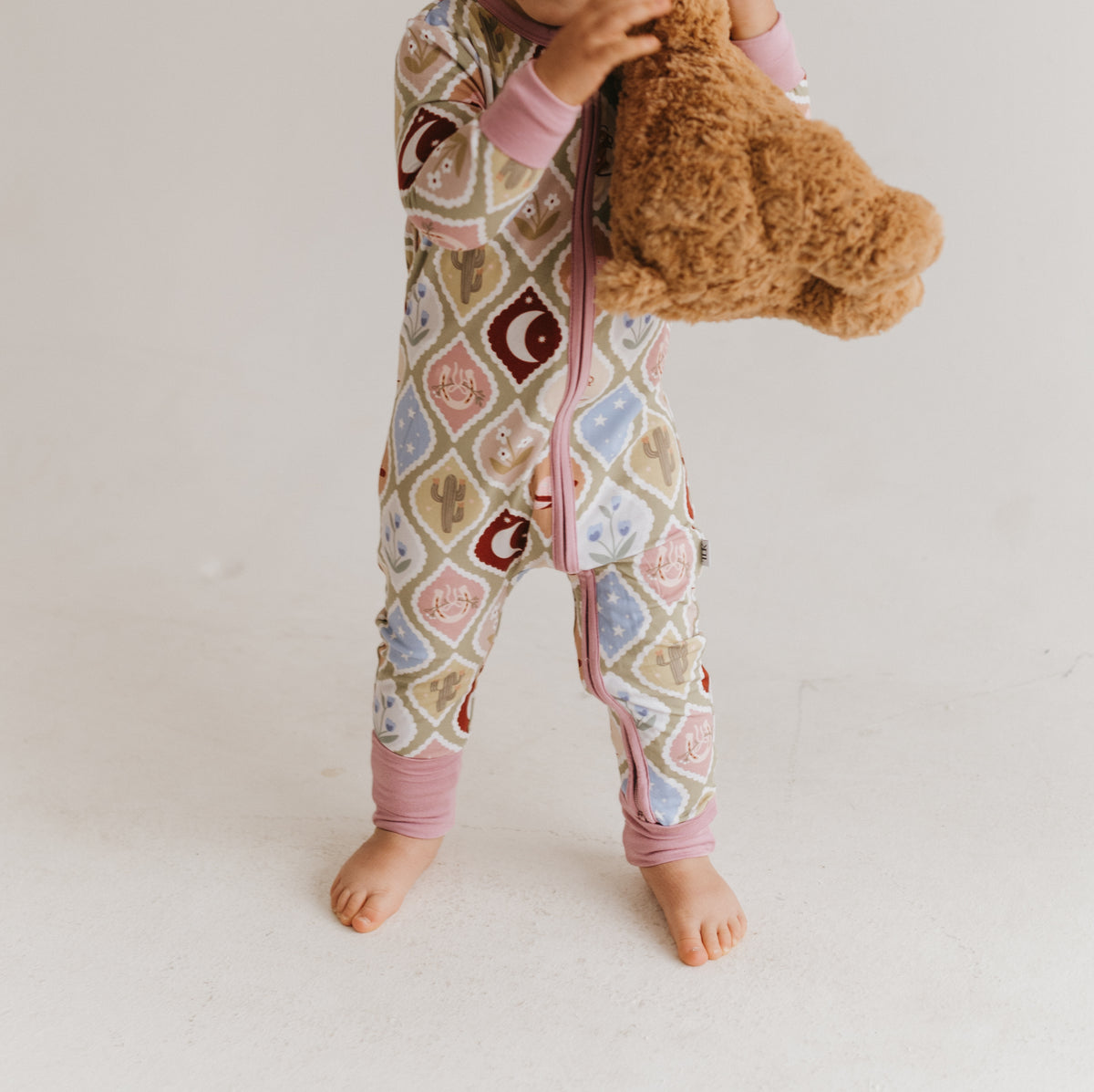 A baby wearing a pink country-themed patterned zip-up sleeper, holding a brown teddy bear against a plain off-white background.