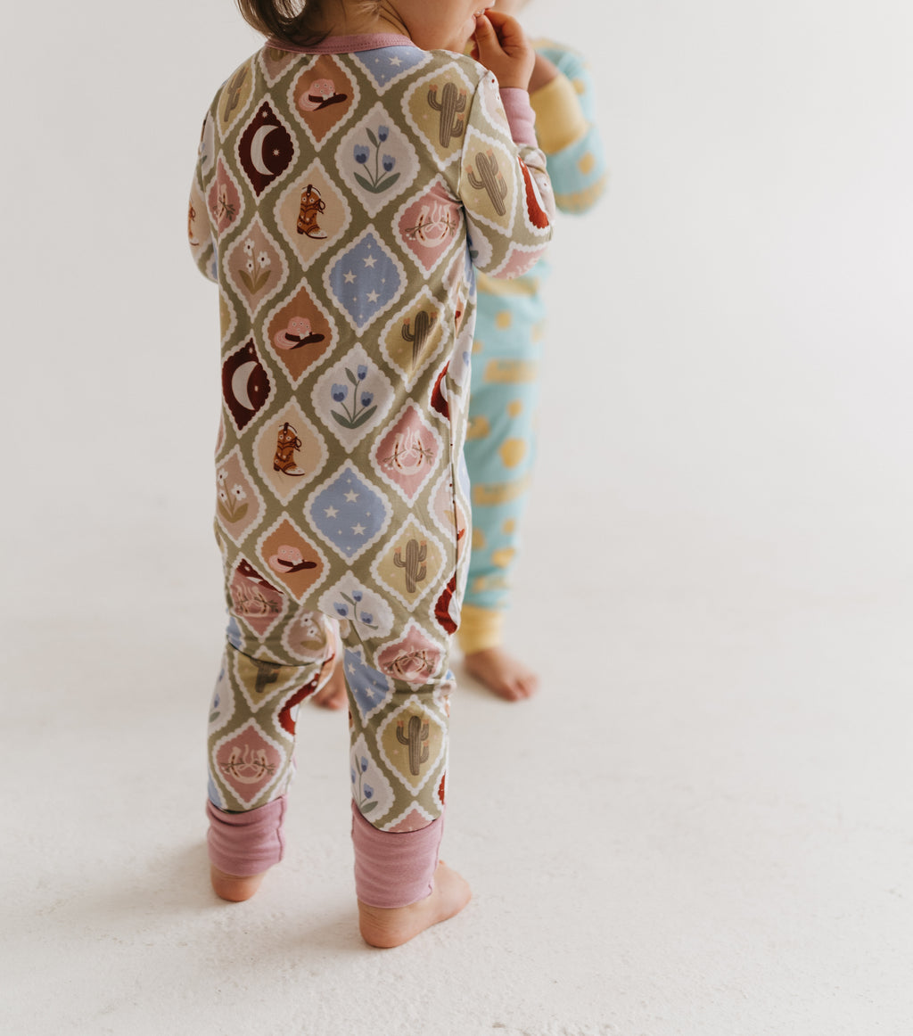 A toddler standing barefoot in a cream-colored patterned one-piece pajama romper with a desert-themed diamond lattice design, with another child partially visible in the background.
