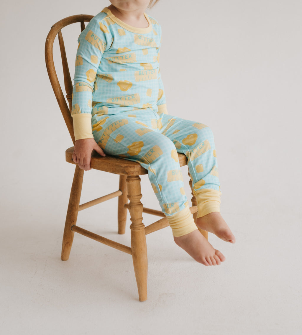 Child sitting on a small wooden chair wearing two piece pajamas set with a yellow and blue butter pattern against a plain white studio background.