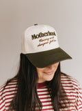A woman wearing a white and dark green trucker-style baseball cap with embroidered text reading 'Motherhood Raising world changers full time' on a plain light gray background.