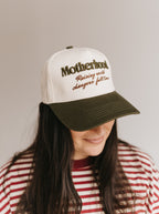 A woman wearing a white and dark green trucker-style baseball cap with embroidered text reading 'Motherhood Raising world changers full time' on a plain light gray background.