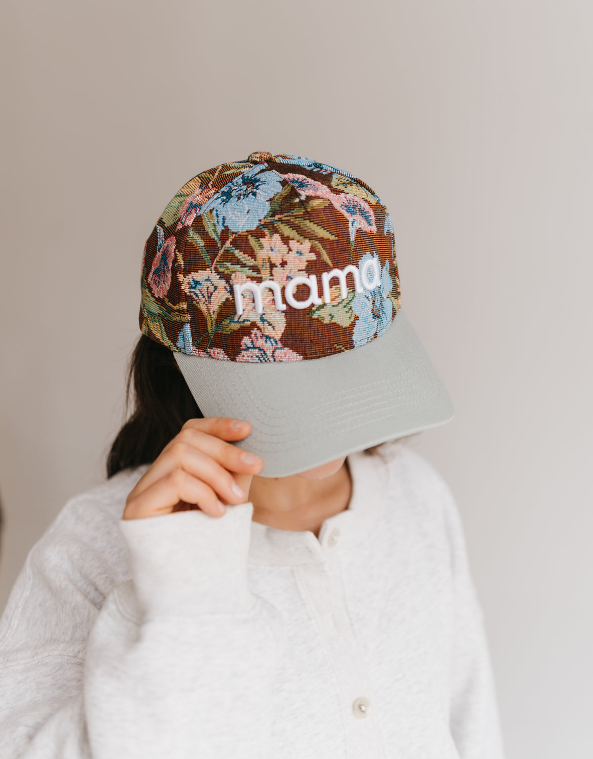 A person wearing a floral patterned baseball cap with a light gray visor and the word 'mama' embroidered on the front, paired with a white knit top against a plain light gray background.