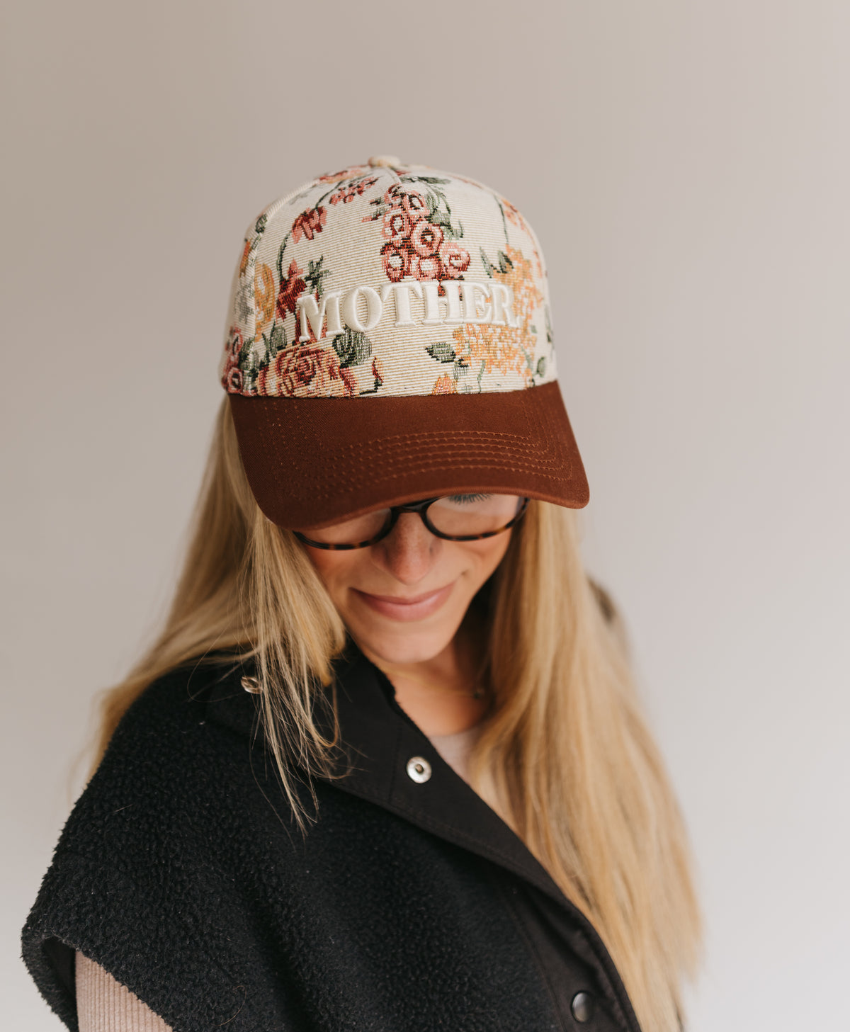 A woman wearing a floral baseball cap with 'MOTHER' embroidered on the front and a brown visor, paired with a black fleece vest against a plain light gray background.