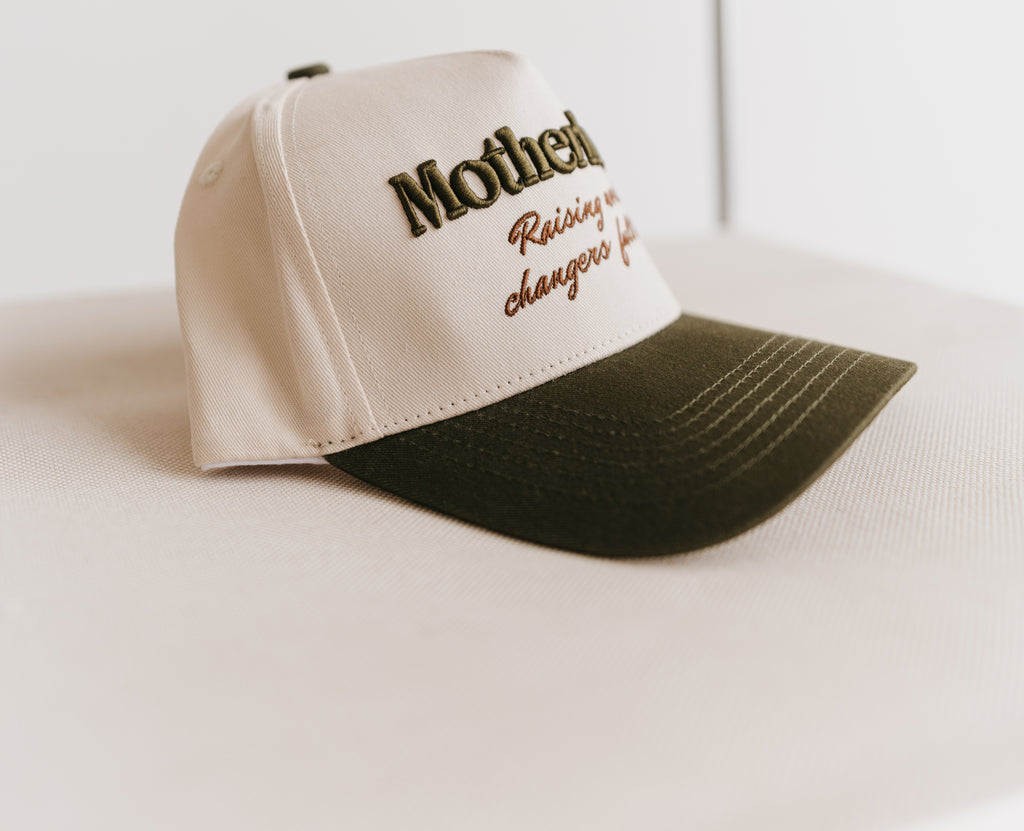 A two-tone baseball cap with a cream-colored front panel and a dark olive green brim, featuring embroidered text on the front that reads 'Motherhood raising world changers full time'