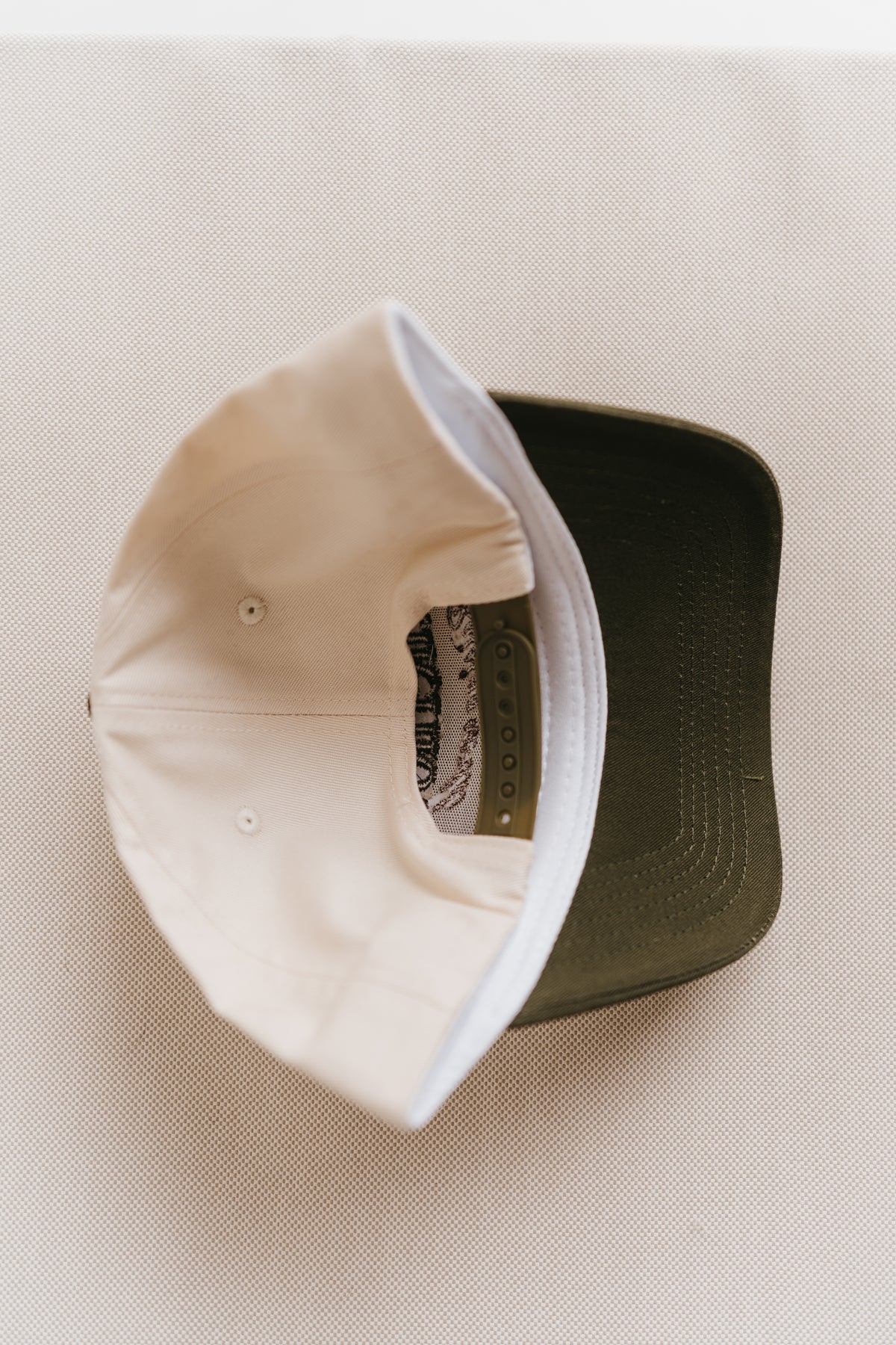 A close-up of a baseball cap shown from the inside, featuring a cream-colored fabric crown and a dark olive green curved brim, laid flat on a textured beige fabric background.