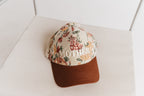 A floral baseball cap with a brown visor, featuring the word 'MOTHER' embroidered in white across the front, resting on a textured white surface.