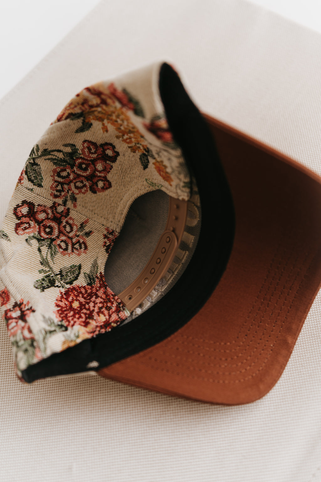 A close-up of a baseball cap with a beige floral patterned fabric crown, a black visor underside, and a brown structured brim, resting on a beige textured surface.