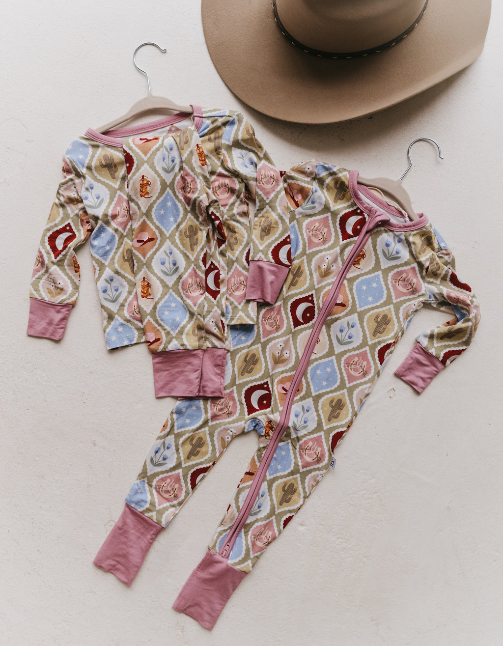 Two pairs on children's long-sleeve pajamas, one two piece and one zip-up, with a colorful geometric pattern, displayed on hangers on a textured off-white background with a cowboy hat partially visible at the top.