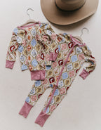 Two pairs on children's long-sleeve pajamas, one two piece and one zip-up, with a colorful geometric pattern, displayed on hangers on a textured off-white background with a cowboy hat partially visible at the top.